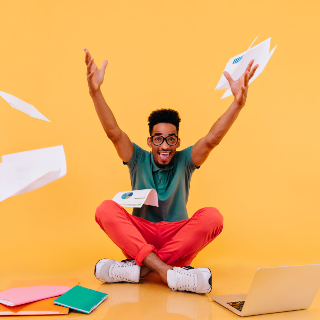 Enthusiastic african student in red pants fooling around during study. Studio shot of smiling male freelancer waving hands on yellow background.